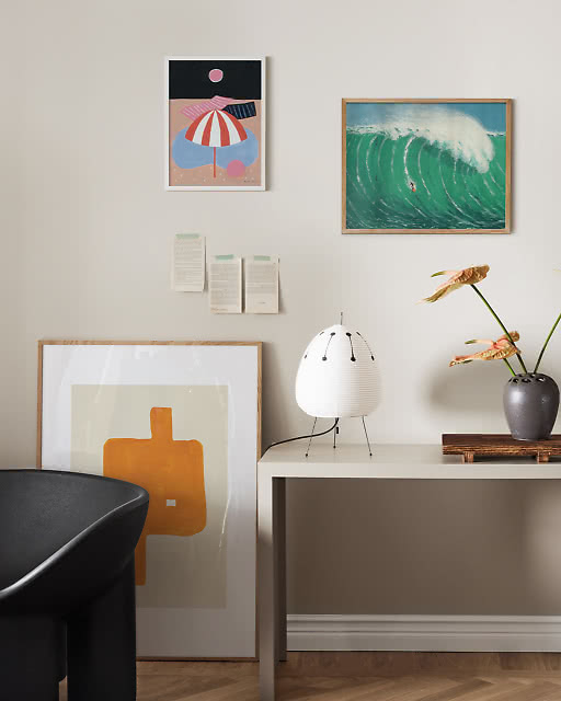 A beach umbrella night scene and a vivid green surfing wave print on a wall, with an orange abstract print leaning below and a paper lantern lamp.